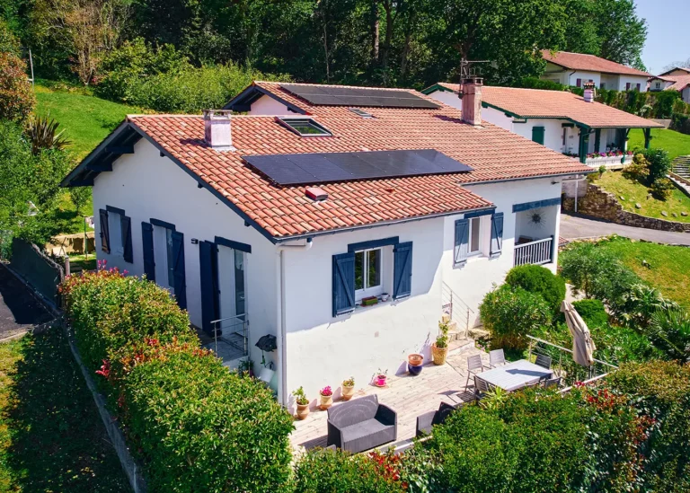 Maison blanche aux volets bleus avec panneaux solaires noirs sur toit en tuiles rouges, vue aérienne sur la terrasse et le jardin arboré.