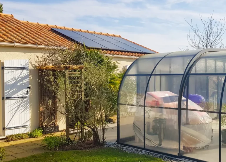 Maison avec toit en tuiles équipée de panneaux solaires noirs, vue sur le jardin et un abri de piscine transparent au premier plan.