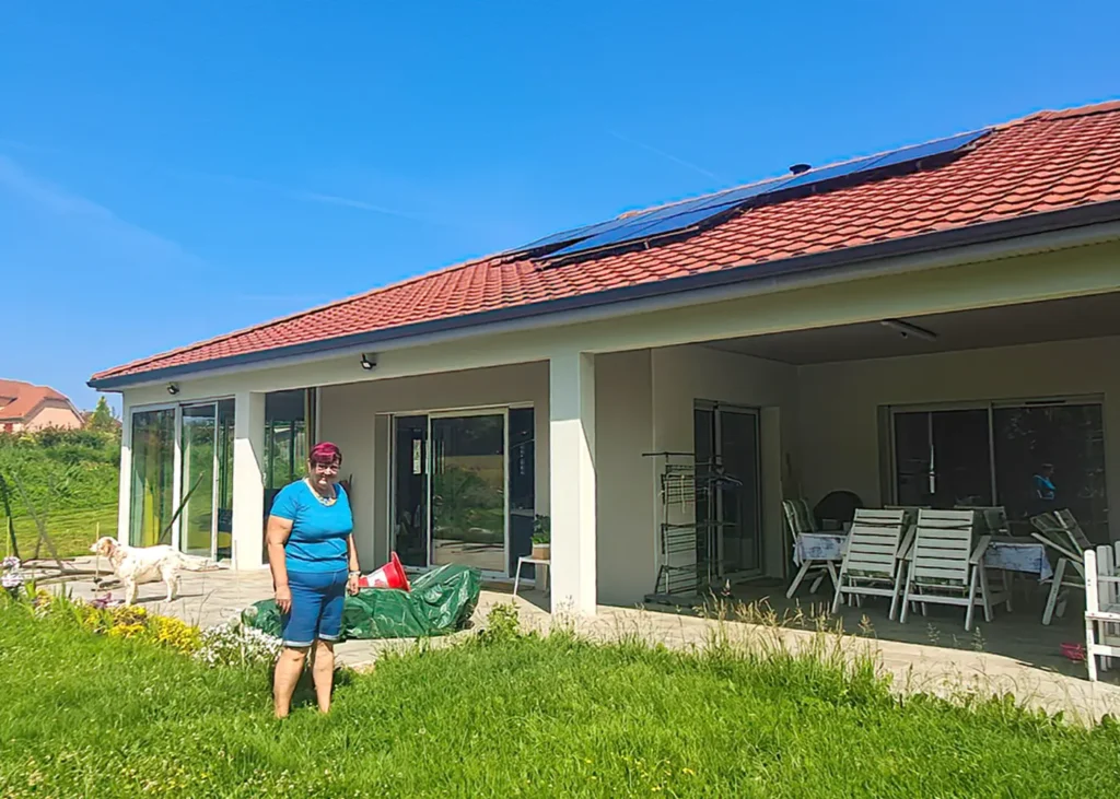 Maison de plain-pied avec toiture en tuiles rouges équipée de panneaux solaires, grande terrasse couverte et personne debout dans le jardin avec un chien en arrière-plan.