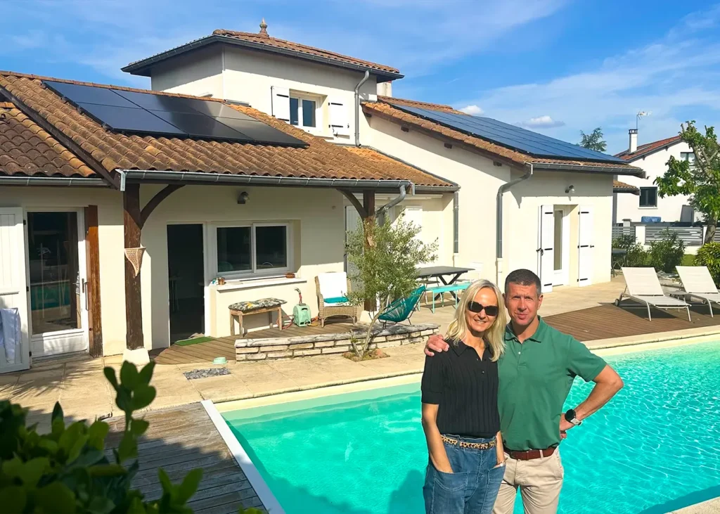 Couple souriant debout au bord de leur piscine devant une maison claire équipée de panneaux solaires noirs sur toit en tuiles.