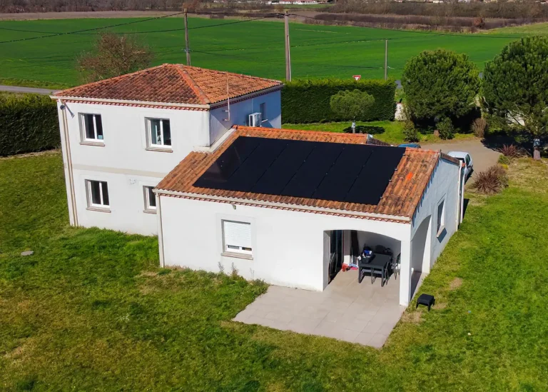 Vue aérienne d’une maison blanche avec toiture en tuiles équipée de panneaux photovoltaïques noirs, entourée de champs et d’un grand jardin.