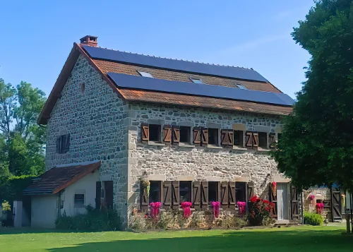 Maison en pierre avec volets en bois, équipée de panneaux photovoltaïques noirs sur la toiture, entourée d’un jardin.